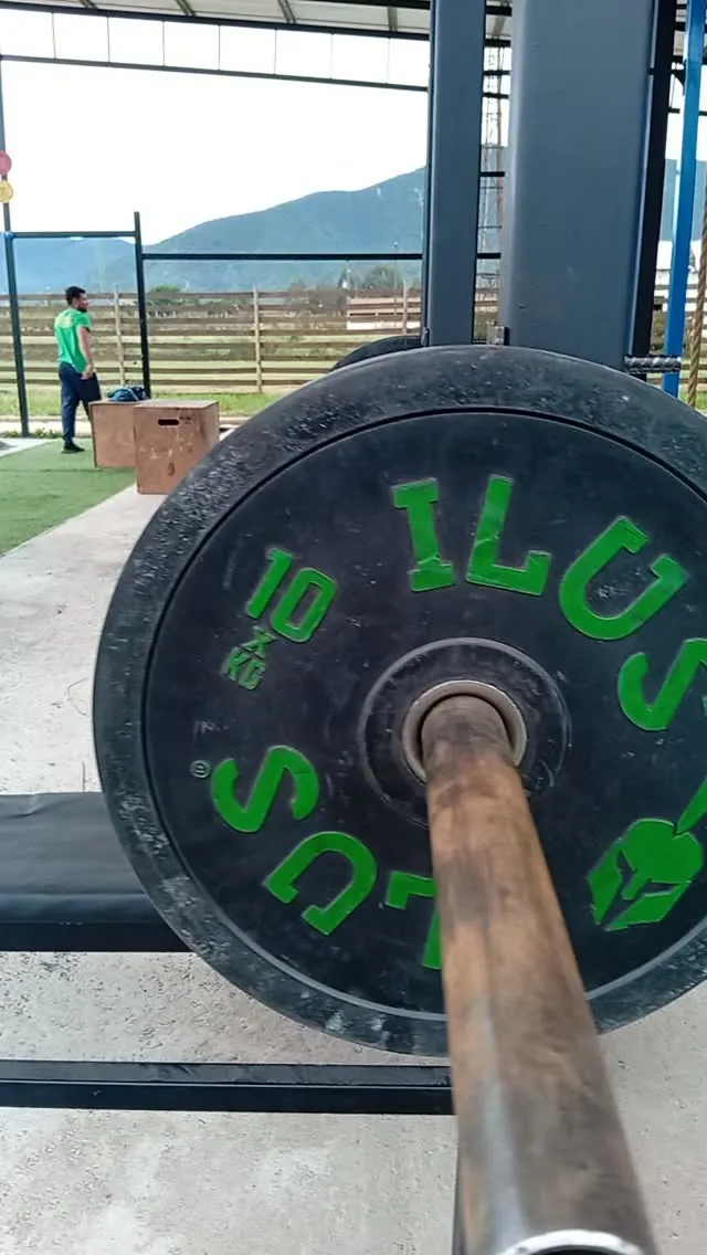 Barra olímpica con un disco de 10 kilos montada en el rack del box, con el pasto y un alumno al fondo